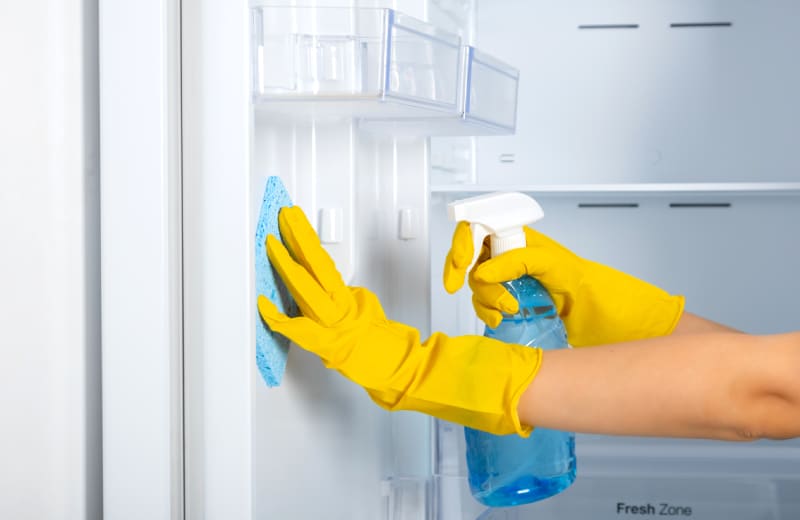 woman's hands in a yellow rubber protective glove and a blue sponge washes, cleans refrigerator shelves